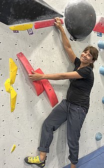 Eine Frau hängt an einer Boulderwand und lacht in die Kamera