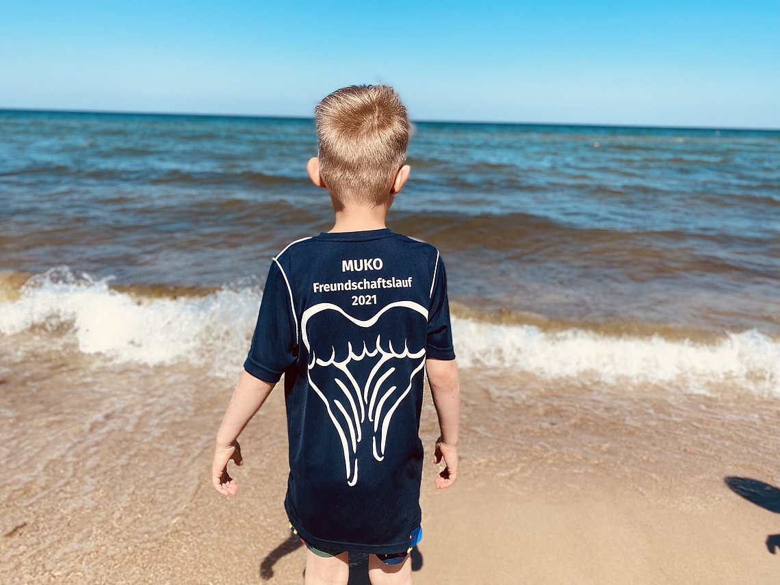 Ein Junge steht am Strand und blickt aufs Meer. Wir sehen ihn von hinten, er trägt ein blaues Trikot, auf dem "MUKO Freundschaftslauf 2021" steht, darunter eine Illustration von Flügeln