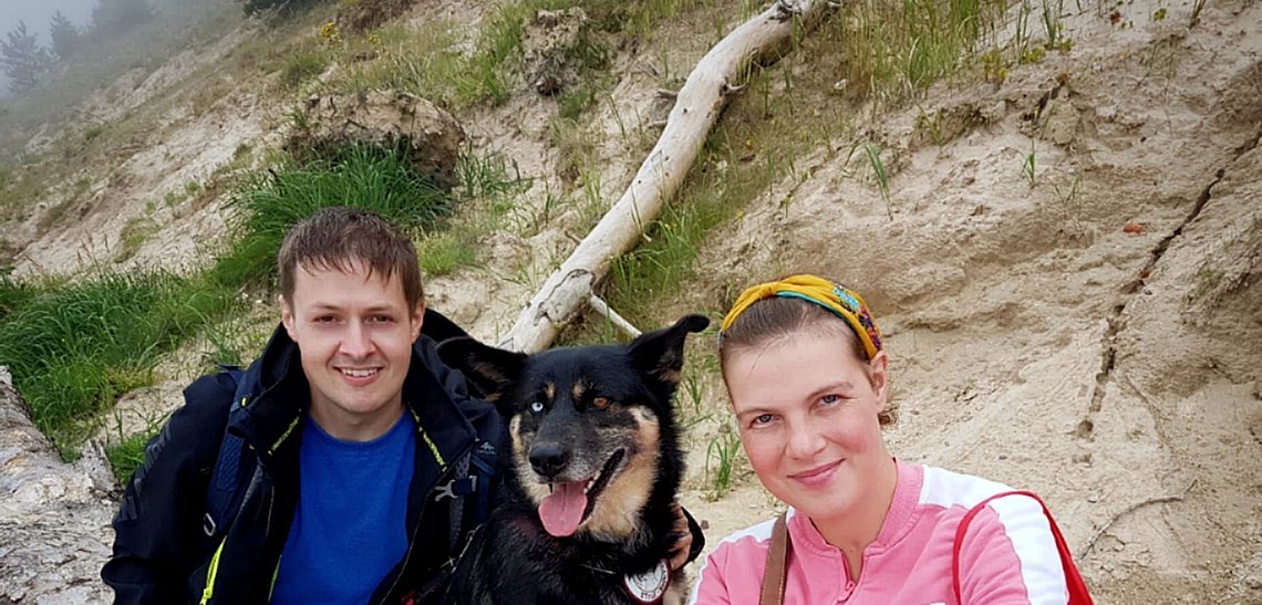 Vor einer Düne hocken ein Mann, ein Hund und eine Frau. Alle drei lachen in die Kamera.