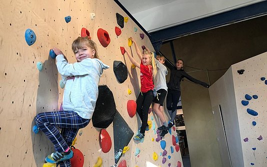 Vier Kinder hängen an einer Boulderwand und lachen in die Kamera
